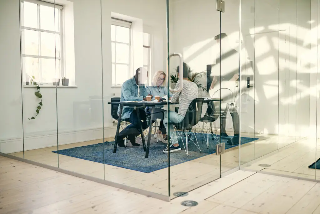 Diverse group of young businesspeople talking together while sitting around a table inside of a glass walled office boardroom